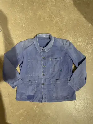 Veste de travail vintage bleue en coton, avec des poches plaquées, une usure authentique et un patch de réparation visible sur une poche.