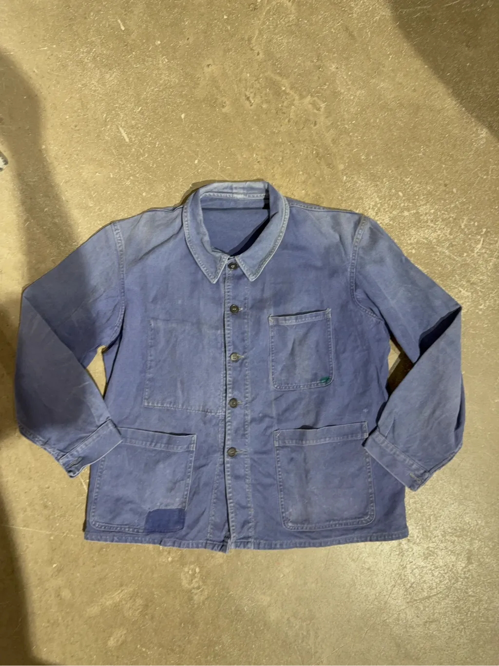 Veste de travail vintage bleue en coton, avec des poches plaquées, une usure authentique et un patch de réparation visible sur une poche.