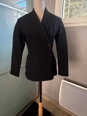 ✨ Une pièce vintage d'exception au style couture et affirmé ! Superbe veste de tailleur / blazer vintage noir de la marque créateur Tony La Marca France, coupe travaillée et très élégante. ✨ Détails & Coupe : Col châle croisé à l'avant, fermeture par bouton, détails de petits boutons décoratifs structurant le côté de la taille. Tissu côtelé / strié de très belle tenue. 📐 Dimensions & Taille : Largeur aisselle à aisselle : 47 cm (convient à un 38 - M). 🧵 Qualité : Belle confection française vintage, pièce originale et structurée.