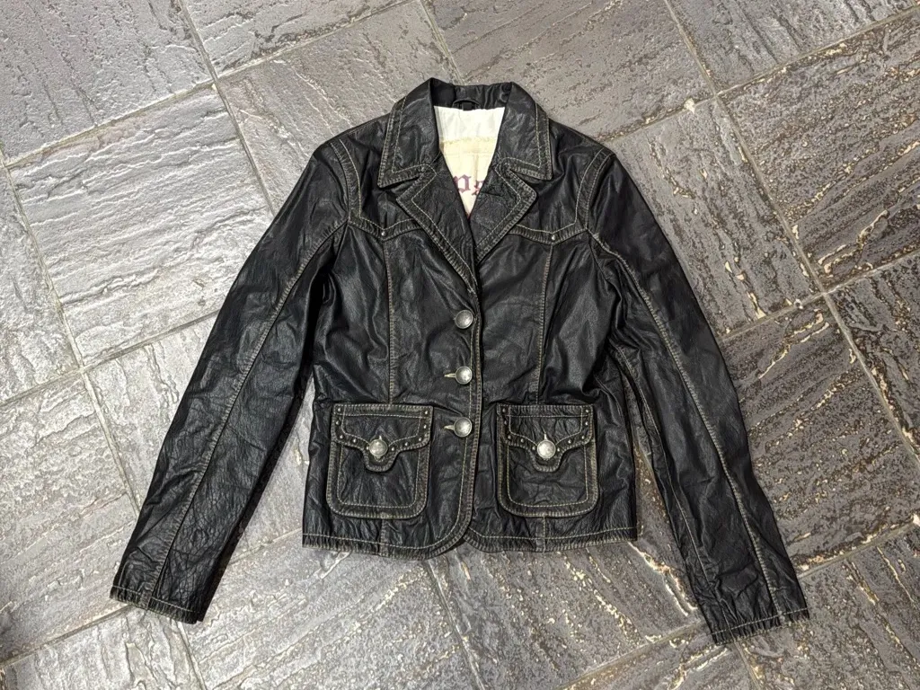 Veste noire en cuir avec des boutons décoratifs argentés et des surpiqûres contrastantes, dotée de deux poches à rabat sur le devant.