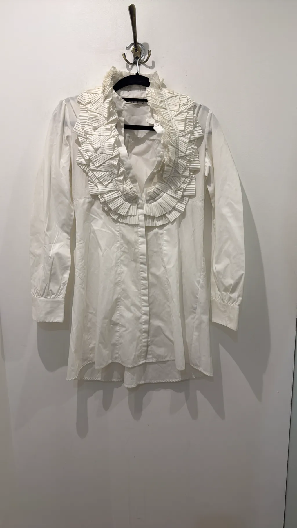 Blouse blanche à manches longues avec un grand col et des volants plissés sur le devant.