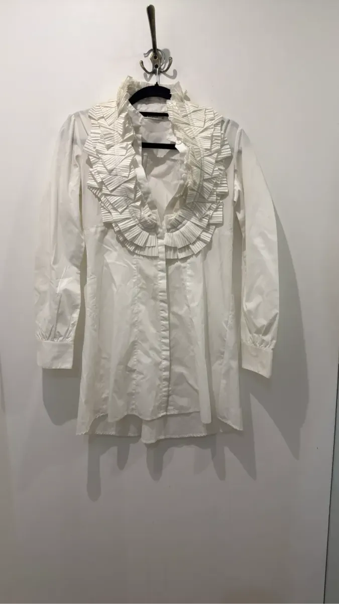 Blouse blanche à manches longues avec un grand col et des volants plissés sur le devant.