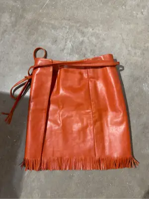 Mini-jupe portefeuille en cuir orange avec franges à l'ourlet.
