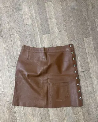 Mini-jupe en cuir marron avec une poche plaquée et des boutons décoratifs sur le côté.